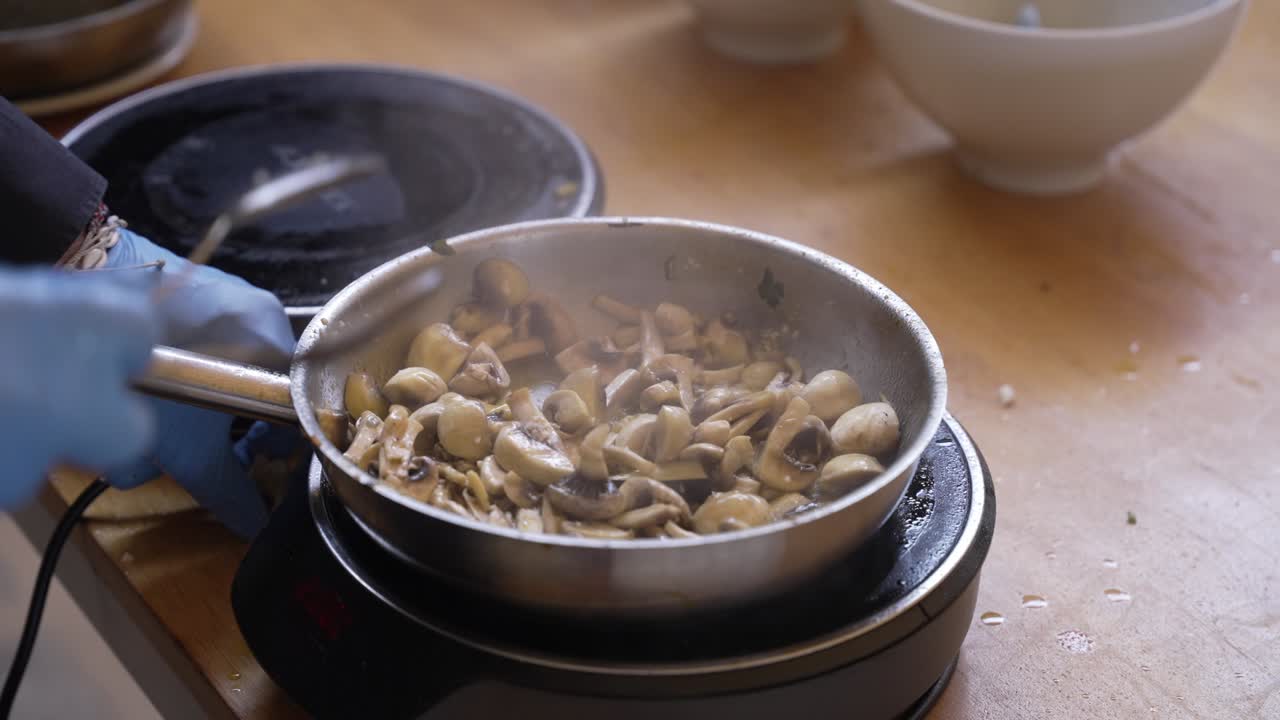 gloved hand stirs mushrooms in sizzling pan on wooden kitchen counter