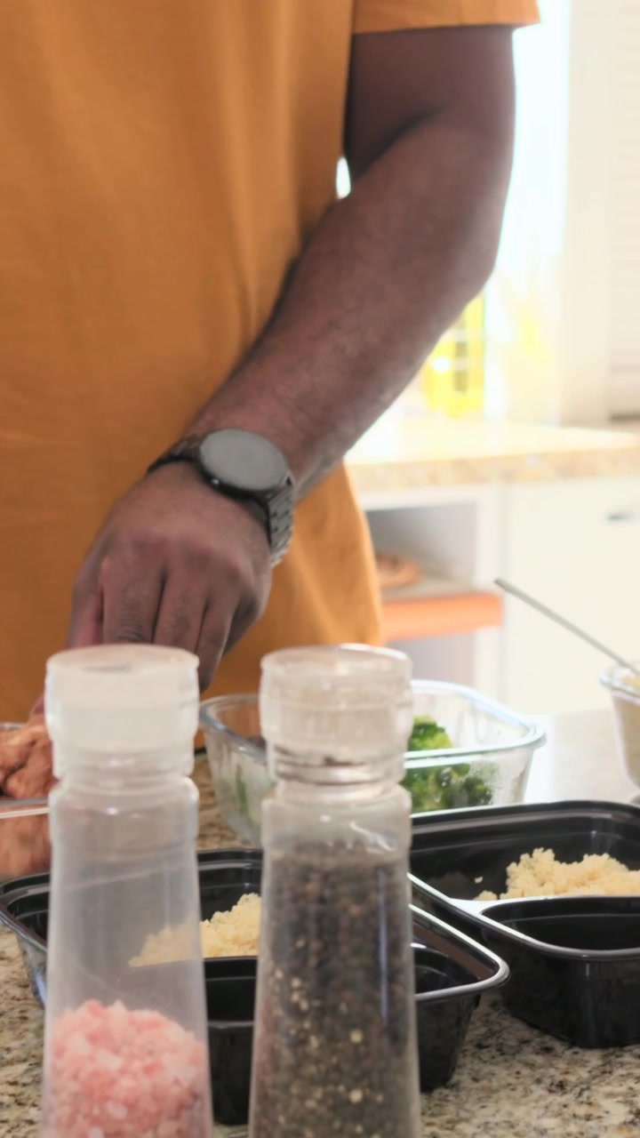 Video verticale: Uomo con maglietta color senape che si allunga per riempire contenitori per pasti con riso, gamberi e broccoli a casa.