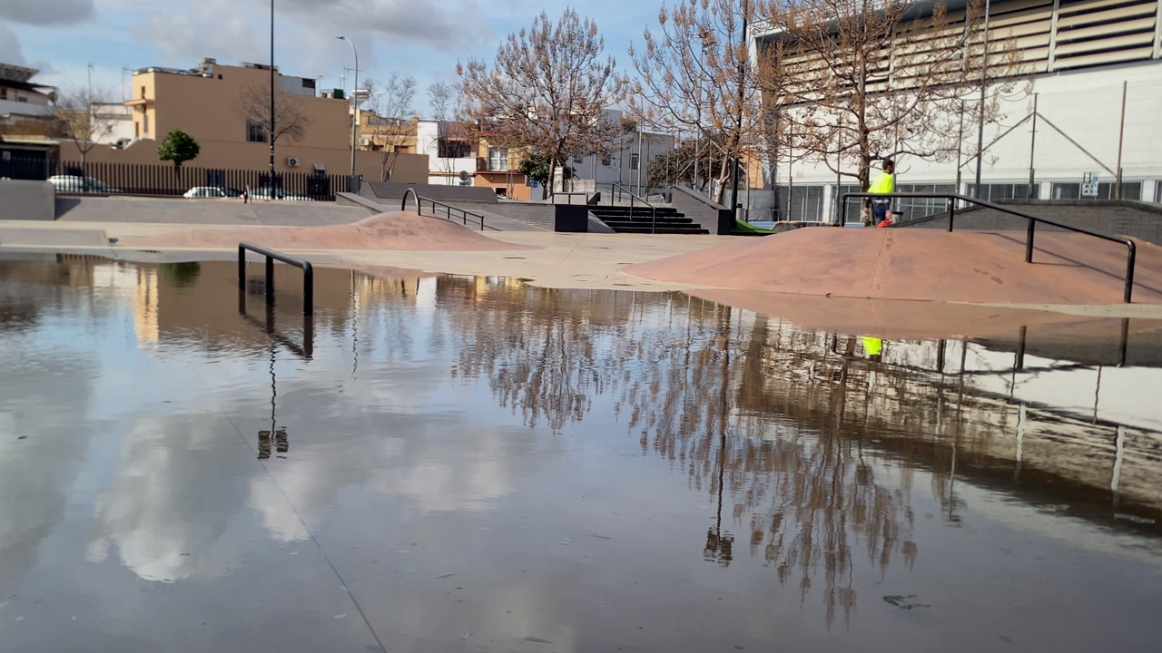 Camas Seville skatepark flooded with rain water. Water reflects bare trees, cloudy sky, urban buildings, and skate ramps