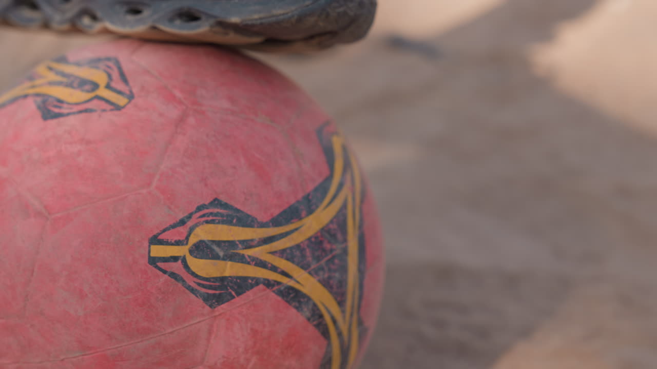 Black sneaker balancing on red ball, worn soccer ball on dusty playground, closeup of scuffed sole pressing into faded panel, focus on balance and ball control, urban street sport vibe with training