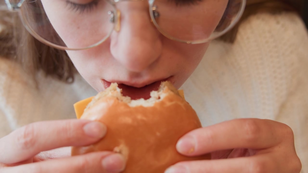 Overhead close-up shows young woman in cream sweater holding bun with both hands, taking bites and chewing as crumbs rest on lips, conveying focused casual snack moment indoors, with woman, eating
