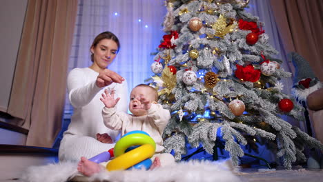 Beautiful Caucasian blond woman sits on the floor with her baby. Mom gives a snowflake decoration from Christmas tree to her son. Low angle view.