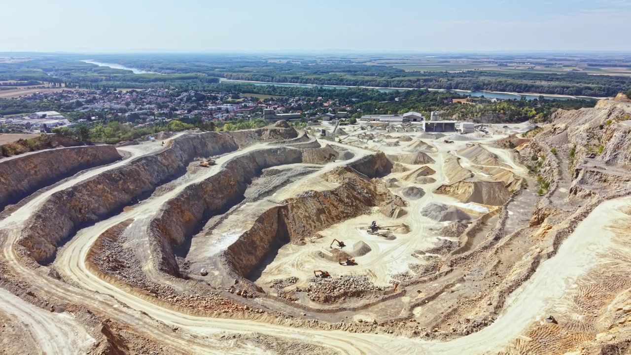 Workers are engaged in quarry activities on a large site with machinery and vehicles near a town and river in the background on a clear day