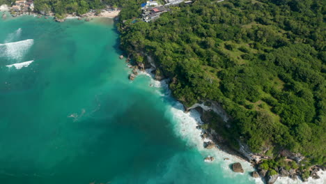 Aerial-view-of-waves-crashing-into-rocky-cliff-on-the-Bali-coastline.-Luxury-resorts-on-lush-tropical-coastline-by-a-turquoise-blue-sea