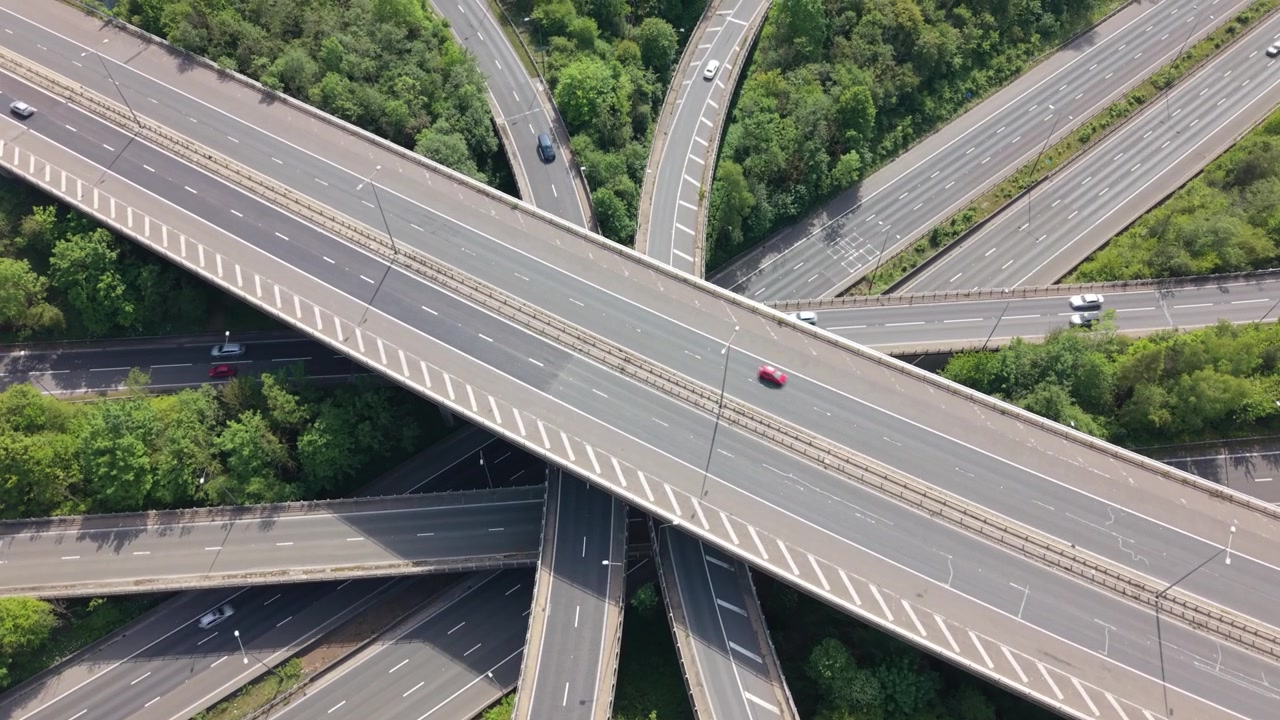High aerial perspective of a complex multilevel highway interchange. Vehicles travel on numerous roads and ramps, surrounded by lush green trees under bright daylight.