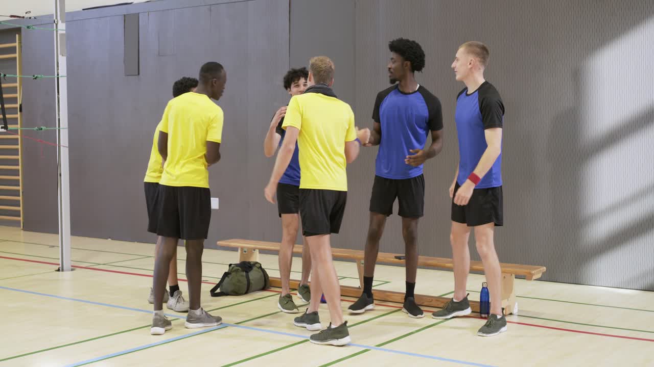 Jugadores masculinos diversos reuniéndose en un banco del gimnasio después de que un jugador con camiseta amarilla hiciera malabares con el balón de voleibol, planificando la jugada.