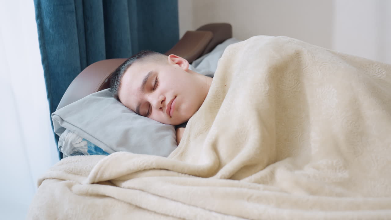 Handsome kid sleeping peacefully under blanket on bed with soft sunlight streaming through window and blue curtain creating warm cozy serene atmosphere of childhood rest calmness comfort