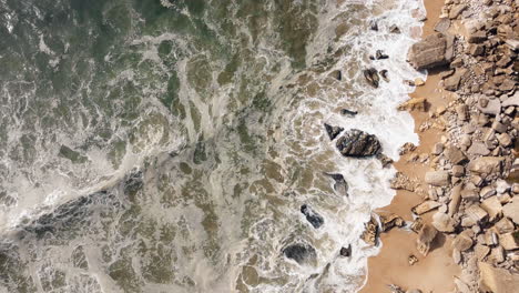 aerial top down shot of crashing waves arriving sand and rocks of nazare beach in summer - circling shot