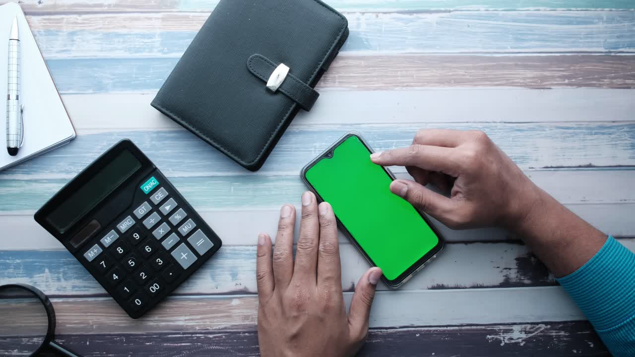 Person using a smartphone with a green screen on a wooden table with other office supplies