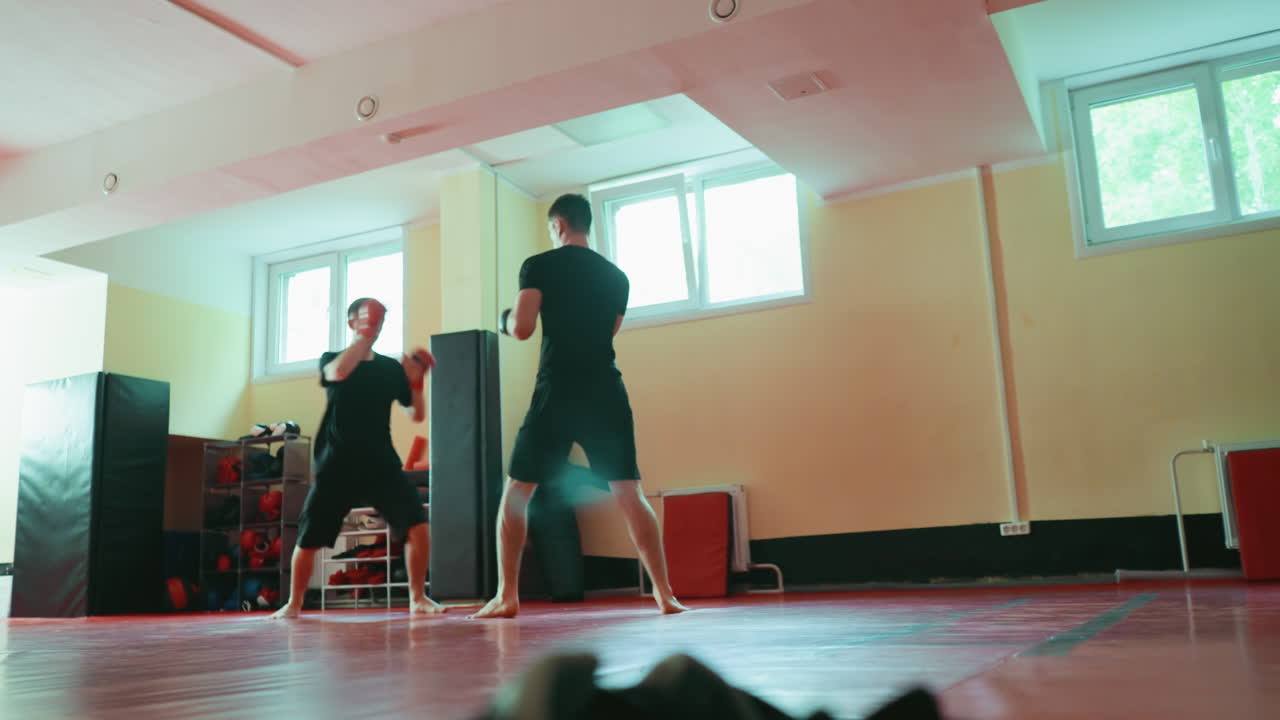 Two fighters sparring in gym on red mat, barefoot, wearing gloves, practicing martial arts with energy and movement, focusing on speed, coordination, and combat training under natural light from windows