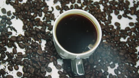 Rotating-cup-of-coffee-with-steam,-surrounded-with-brown-roasted-coffee-beans