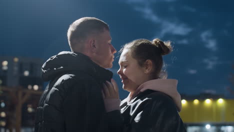 beautiful moment as husband kisses wife on forehead during dusk with eyes closed and soft expressions of love shared under deep sky with warm city lights glowing in background
