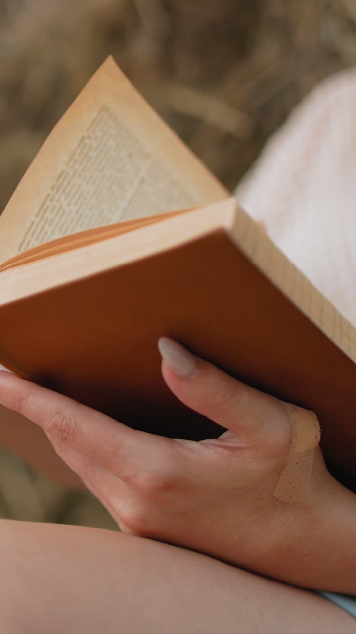 primo piano di una signora che apre una nuova pagina mentre legge all'aperto contro una palla di fieno, le mani tenendo delicatamente il libro, rivelando le pagine allineate, con una morbida sfocatura sullo sfondo