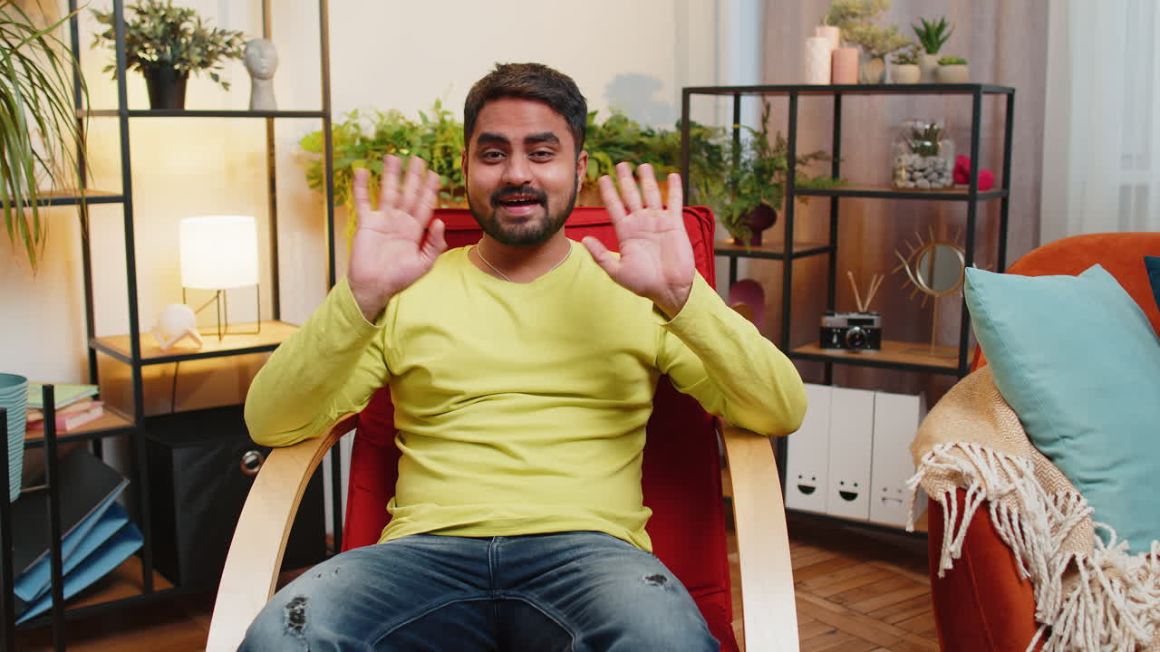 Indian man smiling friendly at camera waving hands gesturing hello hi greeting at home on chair