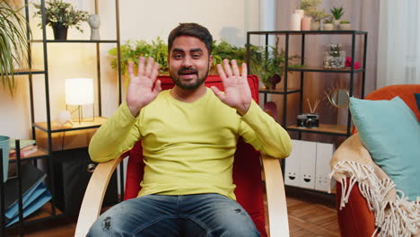 indian man smiling friendly at camera waving hands gesturing hello hi greeting at home on chair