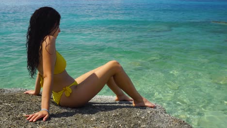 Girl-in-yellow-bikini-meditates-sitting-on-cliff-over-crystal-emerald-water-of-turquoise-sea-in-Ionian-coastline