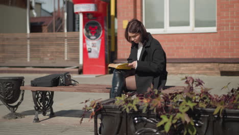 researcher seated outdoors with legs crossed, focusing on the book in her hands. in the background, a machine beeps while flowers in a pot sway, on a sunny day