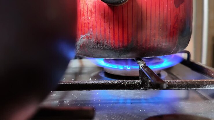 Close-up of a red pot heating over a blue gas burner on a kitchen stove, showing the underside and flame with warm indoor light and shallow depth of field