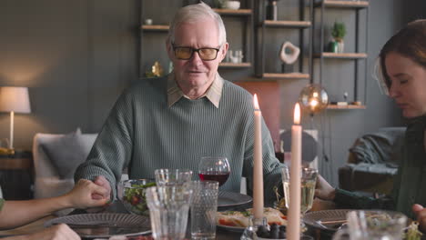 old man and his two adult daughters holding hands and praying before meal while sitting at dinner table at home