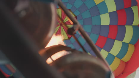 upward view shows powerful flame from metal burner firing into vibrant hot air balloon canopy, highlighting vivid geometric pattern of fabric as balloon inflates and prepares for lift under clear sky