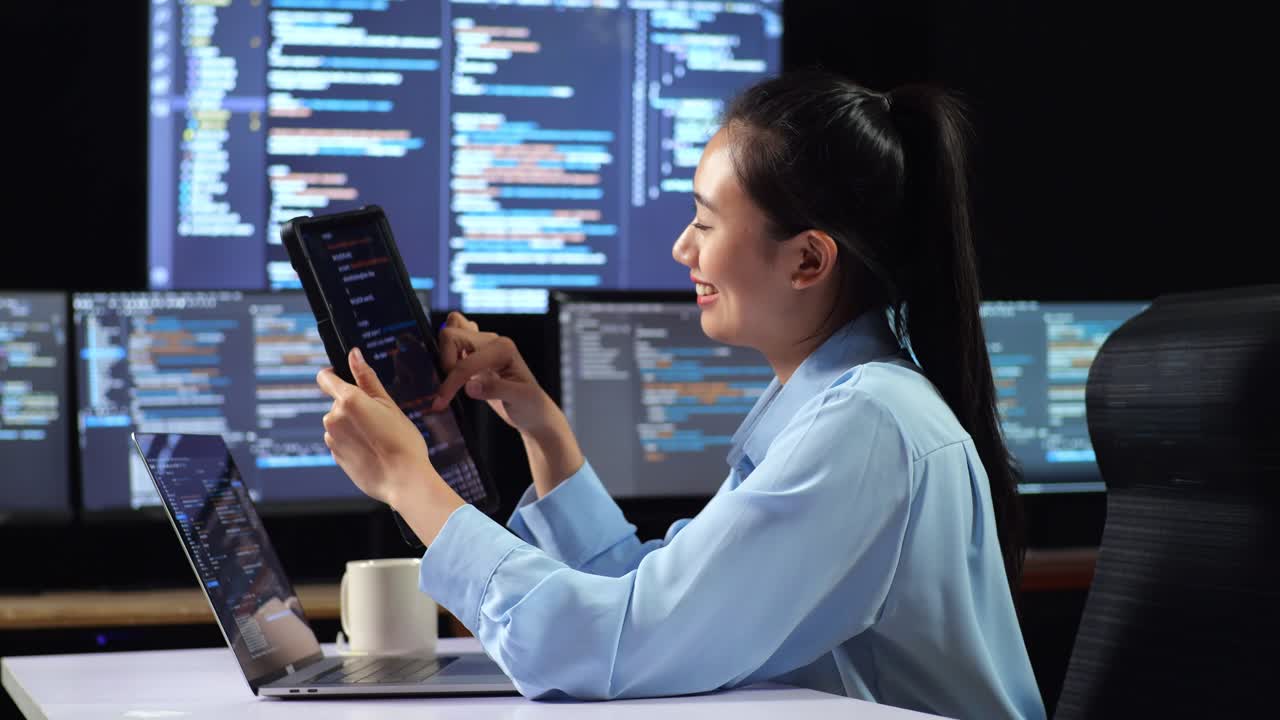 Side View Of Asian Female Programmer Looking At Database On Tablet While Writing Code By A Laptop Using Multiple Monitors Showing Database On Desktops In The Office