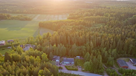 Aerial-view-backwards-over-a-off-grid-house-in-middle-of-sunlit-forest-and-rural-fields