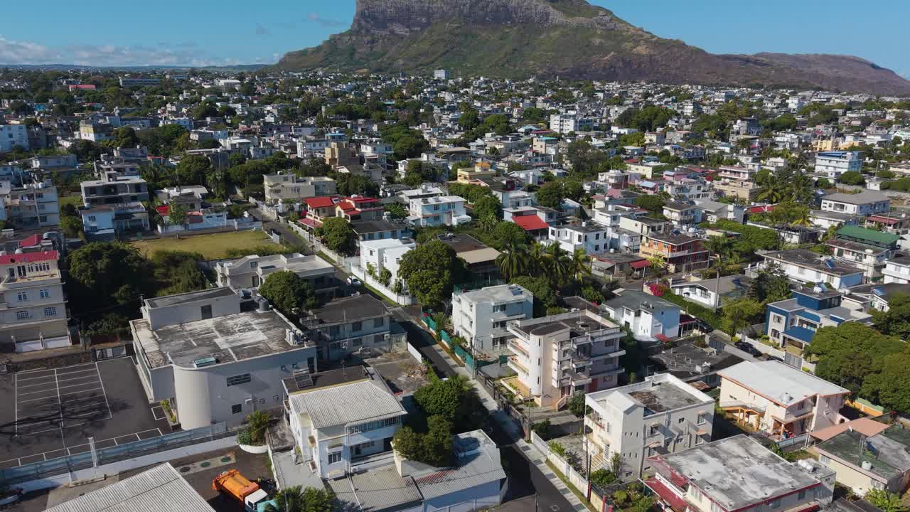 Drone shot of Beau Bassin–Rose Hill, Mauritius, showcasing a tropical suburban landscape with colorful houses, lush greenery, and the majestic mountain range rising in the background under a blue sky