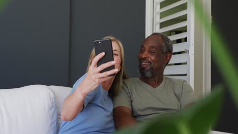 Pareja-De-Ancianos-De-Raza-Mixta-Tomando-Un-Selfie-En-Un-Teléfono-Inteligente-Mientras-Está-Sentado-En-El-Sofá-De-Casa