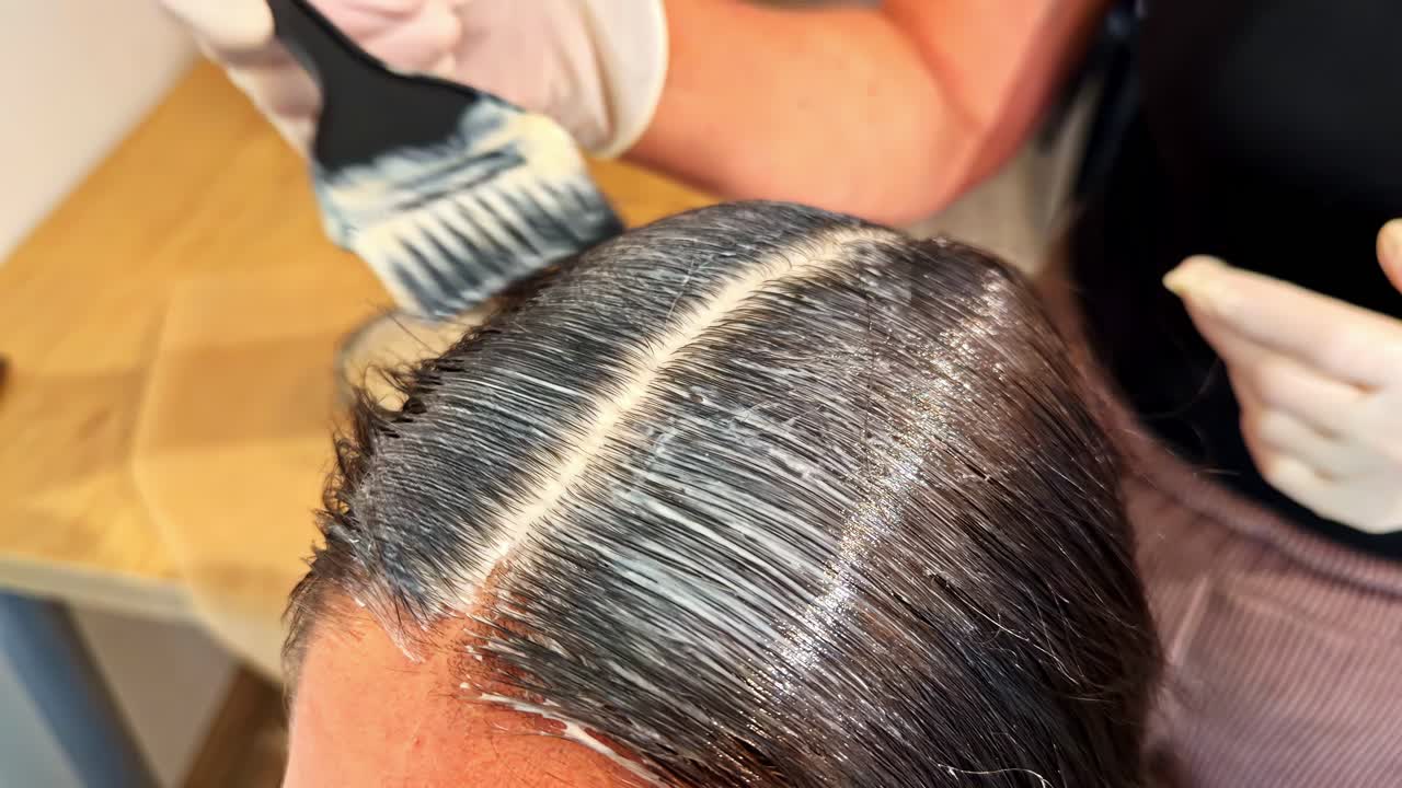 Stylist's Hand In Gloves Applying Hair Dye Onto Dark Hair Using Brush At Salon. closeup shot