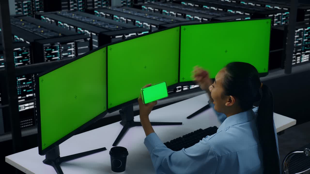 vista laterale di una donna asiatica felice che guarda lo schermo verde del cellulare e celebra mentre lavora con più monitor di computer nel centro dati