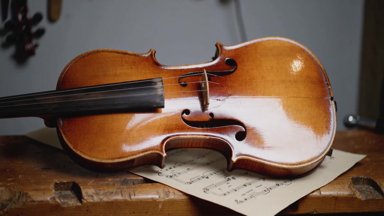 Lumière changeante révélant un violon en bois reposant sur un établi d'atelier usé, la partition de musique devenant plus claire