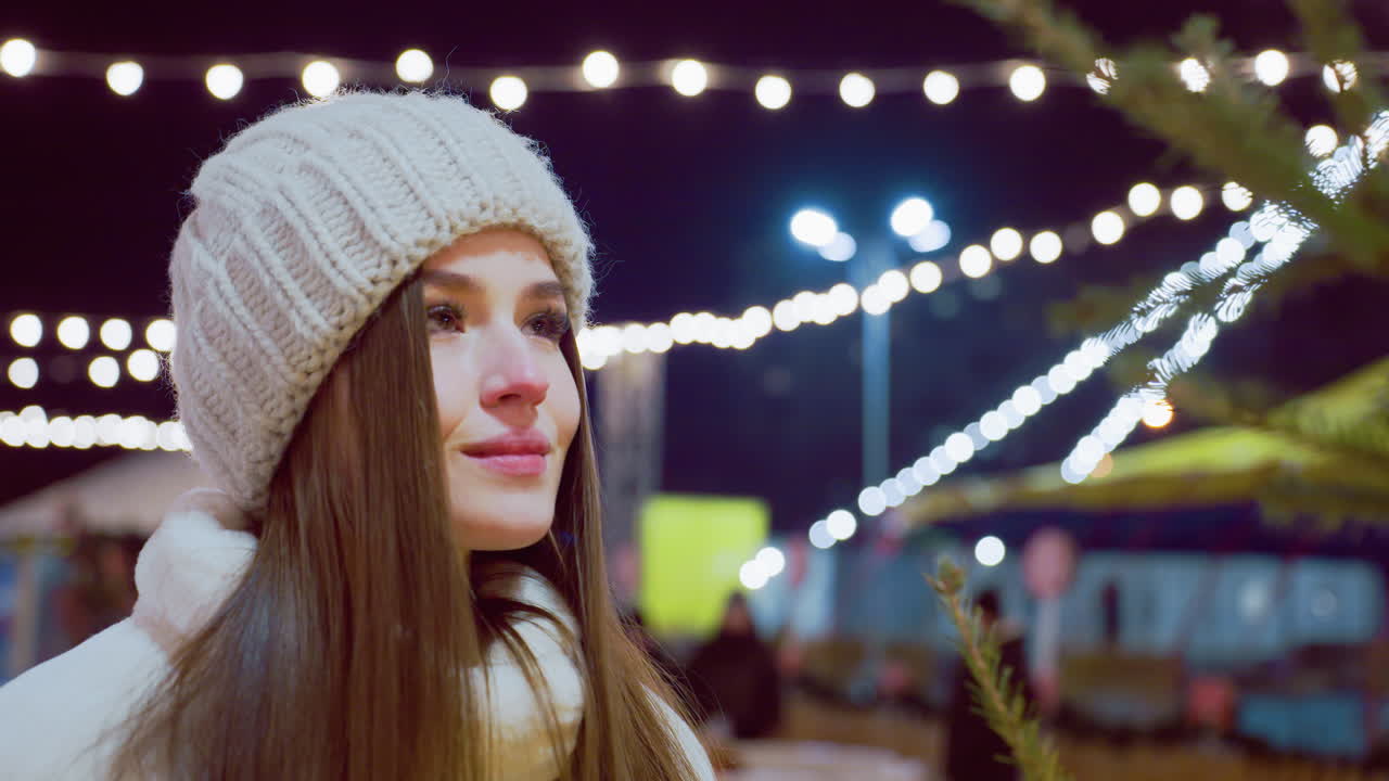 Elegant woman in white beanie and cozy winter coat admiring beautifully decorated pine tree, soft festive lights and blurred people in background