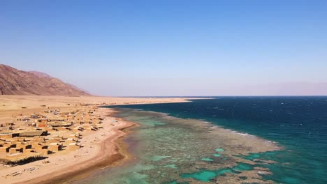 Se-Despliega-Una-Vista-Aérea-De-Dahab-En-Egipto,-Que-Revela-Playas-Bañadas-Por-El-Sol,-Aguas-Cristalinas-Y-Majestuosas-Montañas-Como-Telón-De-Fondo.