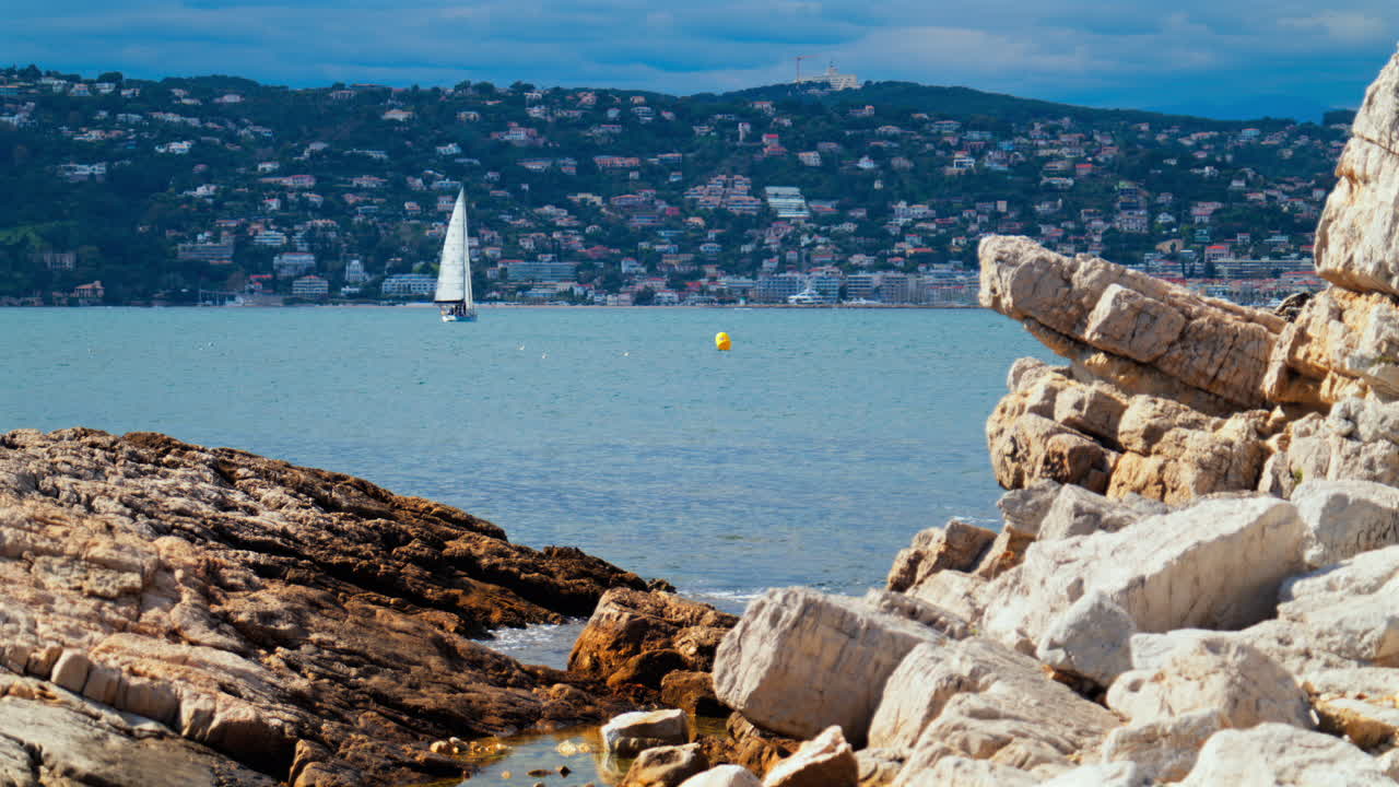 Rocks on the shore wit a sail boat moving on the sea in Juan-les-Pins, France with the city and the mountain on the background
