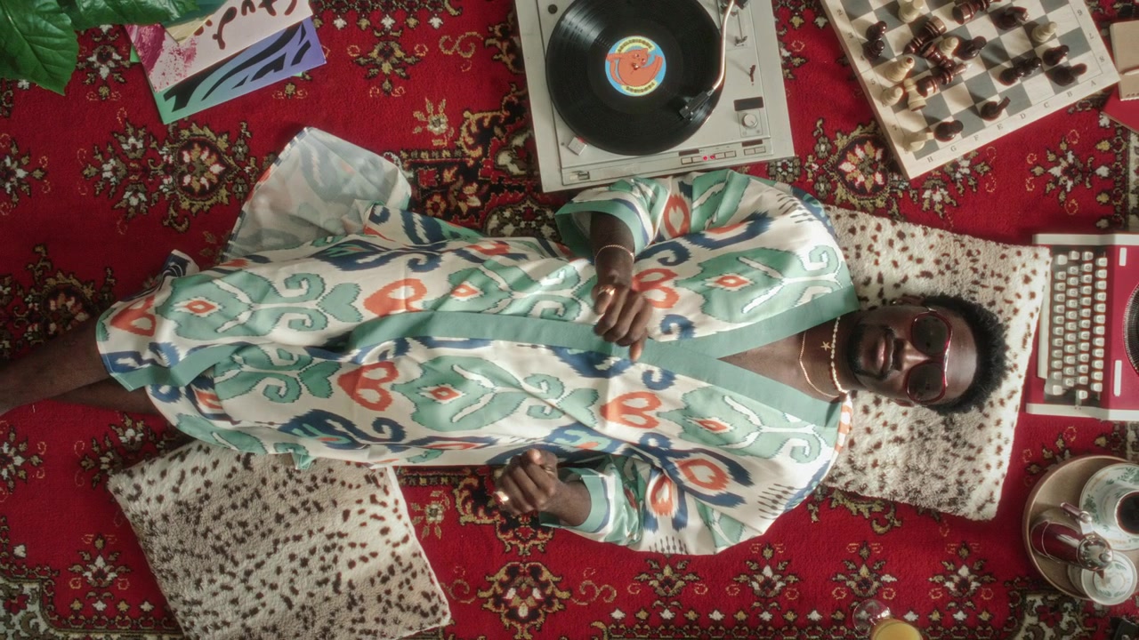 Afslappet sort mand i morgenkåbe danser til vinylmusik, mens han ligger på et vintage tæppe