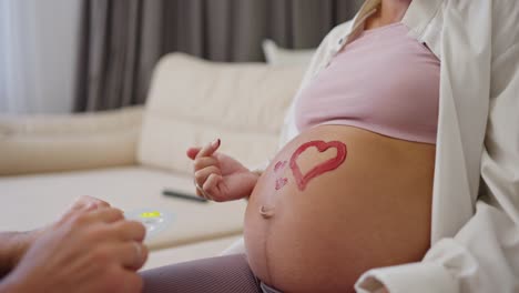 Close-up-a-man-using-red-paint-draws-a-heart-on-the-belly-of-his-pregnant-wife-on-the-sofa-at-home.-Happy-couple-expecting-baby-at-home