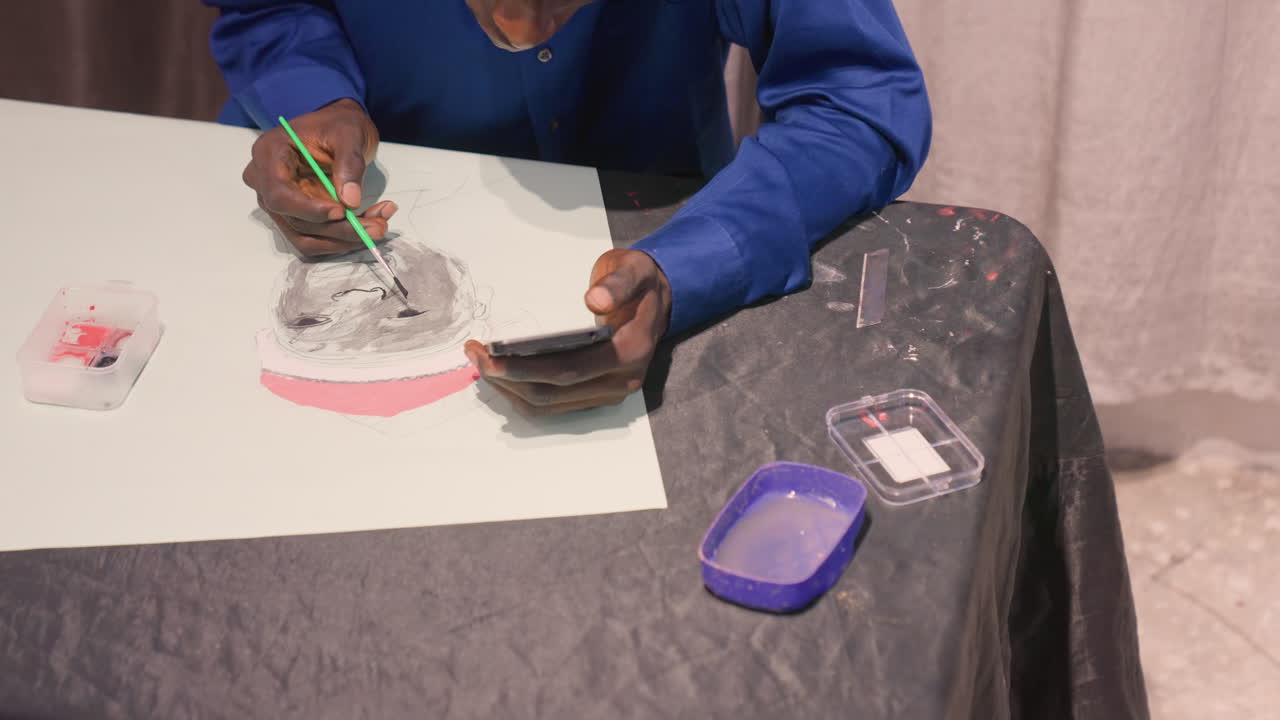 Black artist hands painting portrait study, darkskinned male mixing black ink and gray wash on white paper, green brush in right hand, phone reference in left hand, plastic palette and container
