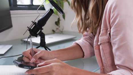 woman recording podcast at home, writing notes and using smartphone on desk