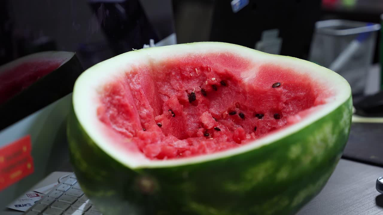Un'anguria fresca con polpa rossa e semi neri appoggiata su una scrivania in legno accanto a una tastiera. Un'opzione di spuntino sano per i dipendenti in un moderno spazio di lavoro interno