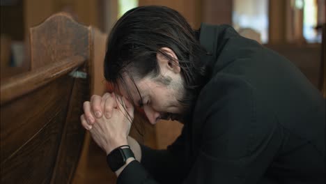 young man in black suit praying in church pew in cinematic slow motion with folded hands