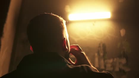 rear shot of man smoking and exhaling cigarette smoke from his mouth in front of light