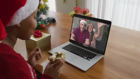 Afroamerikanische-Frau-Mit-Weihnachtsmütze-Nutzt-Laptop-Für-Weihnachtsvideoanruf-Mit-Paar-Auf-Dem-Bildschirm