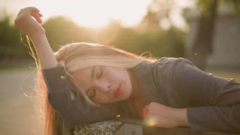 woman seated on bench resting head on arm while gently touching hair, soft sunlight creating warm ambience around her, with a blur background