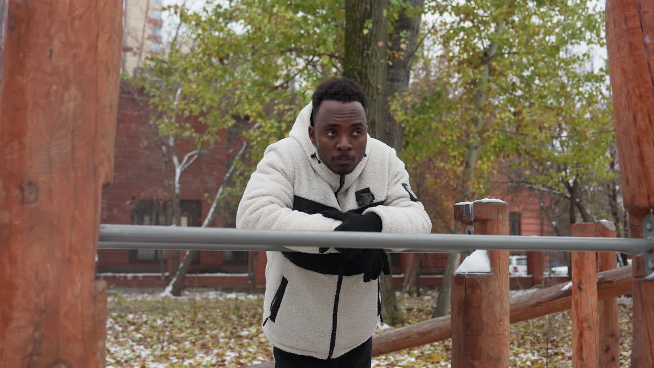 Contemplative young adult leans on iron bar, looking around in deep thought, standing in outdoor workout area with snow-covered ground, residential buildings and trees in background