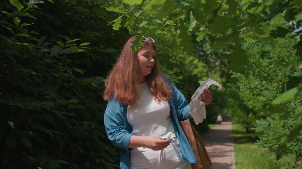 Chubby lady wearing blue shirt and white top smiling joyfully while admiring embroidered fabric in hand and dancing lightly under green leaves, enjoying creative inspiration