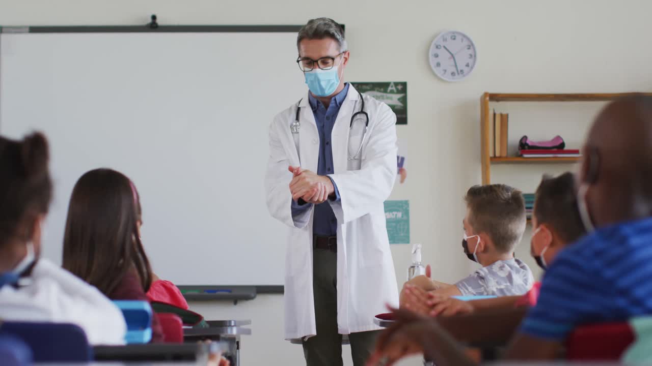 varios maestros desinfectando las manos en el aula, con los escolares sentados con máscaras faciales