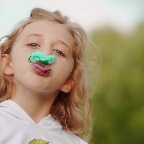 funny girl with a piece of sweet cotton candy on her face. pretty little girl eating green cotton candy on blur nature background.