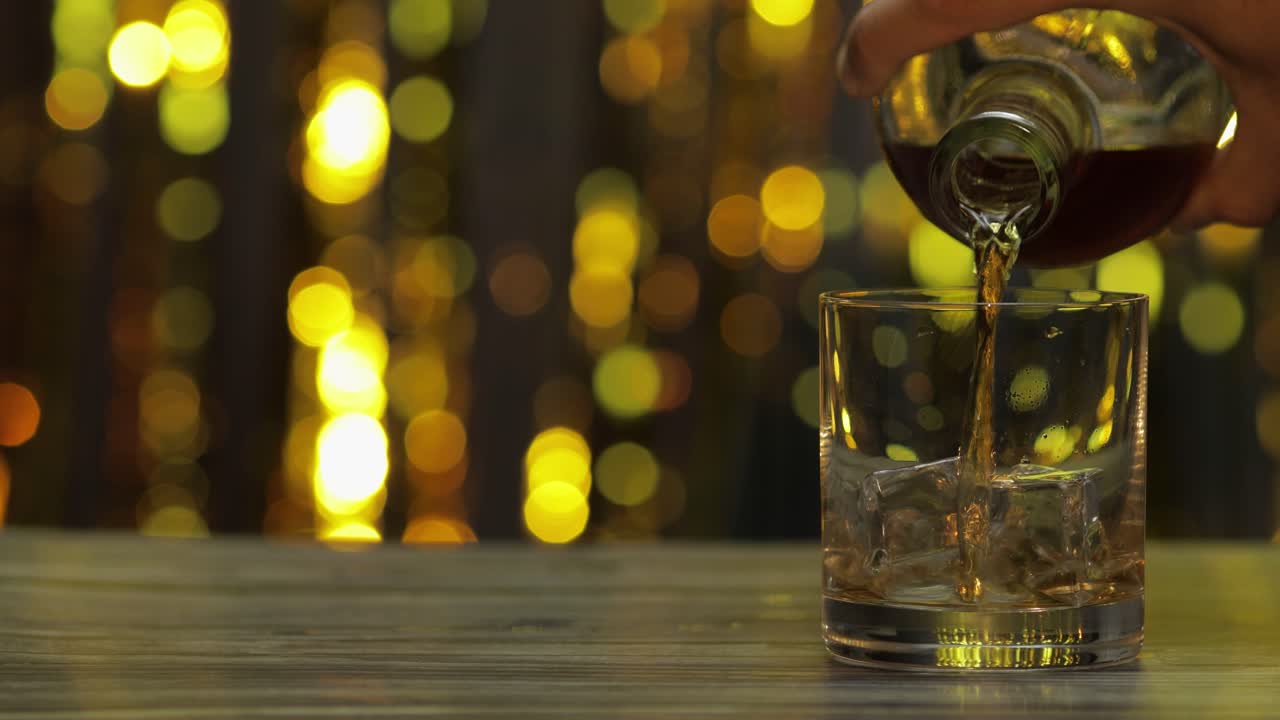 vertido de whisky dorado, coñac o brandy de la botella en un vaso con cubos de hielo. fondo brillante