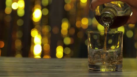 pouring of golden whiskey, cognac or brandy from bottle into glass with ice cubes. shiny background