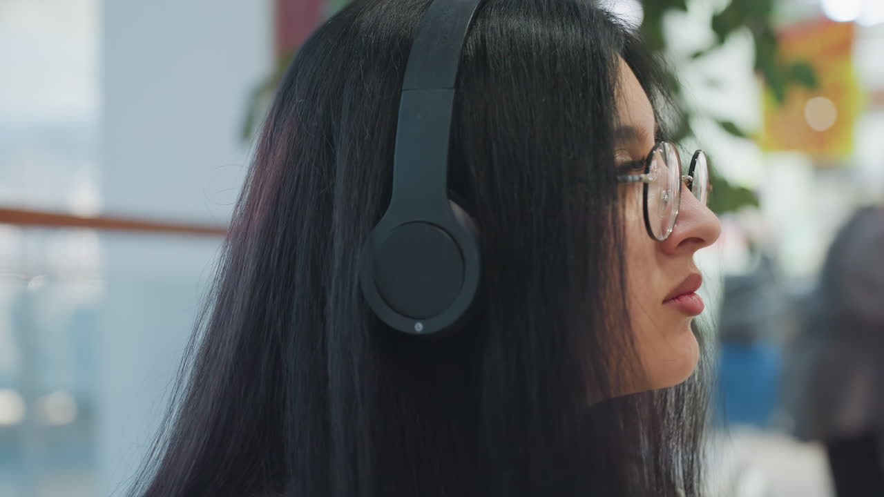 Close up of woman wearing headphones and glasses looking around contemplatively with long dark hair, standing indoors in brightly lit mall with blurred movement of people in background